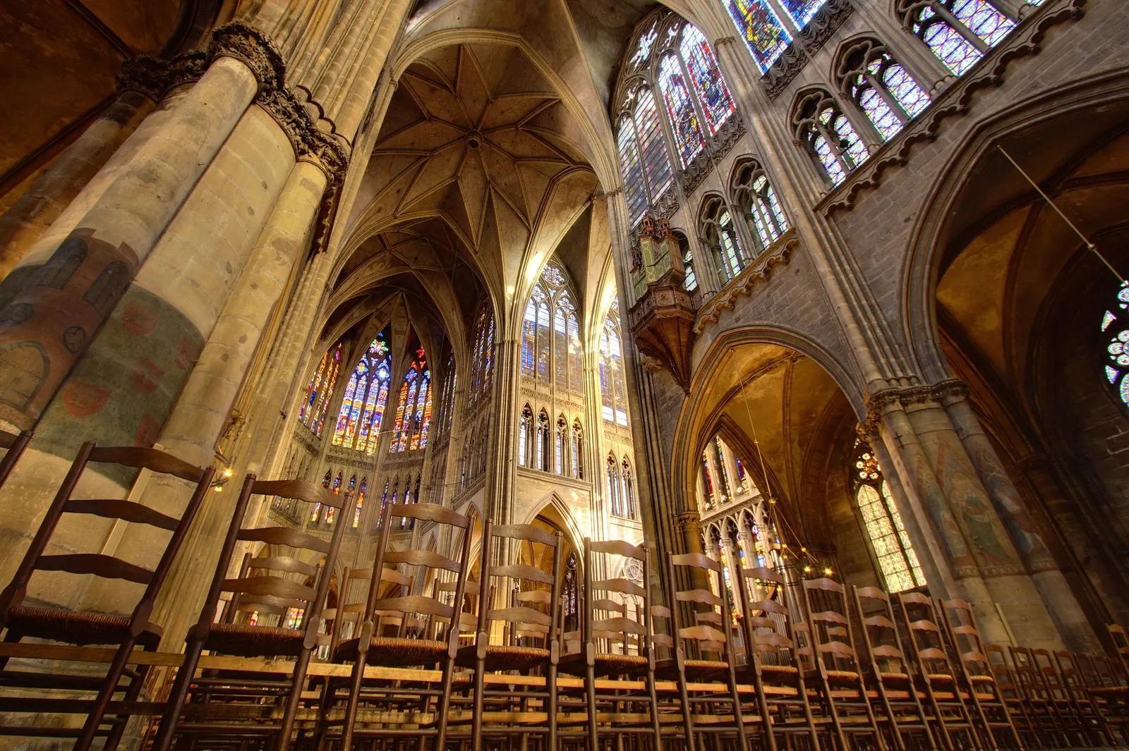 Vue en contre-plongée de l'intérieur de la cathédrale de Metz, piliers, voûtes et grandes verrières colorées au-dessus des rangées de chaises